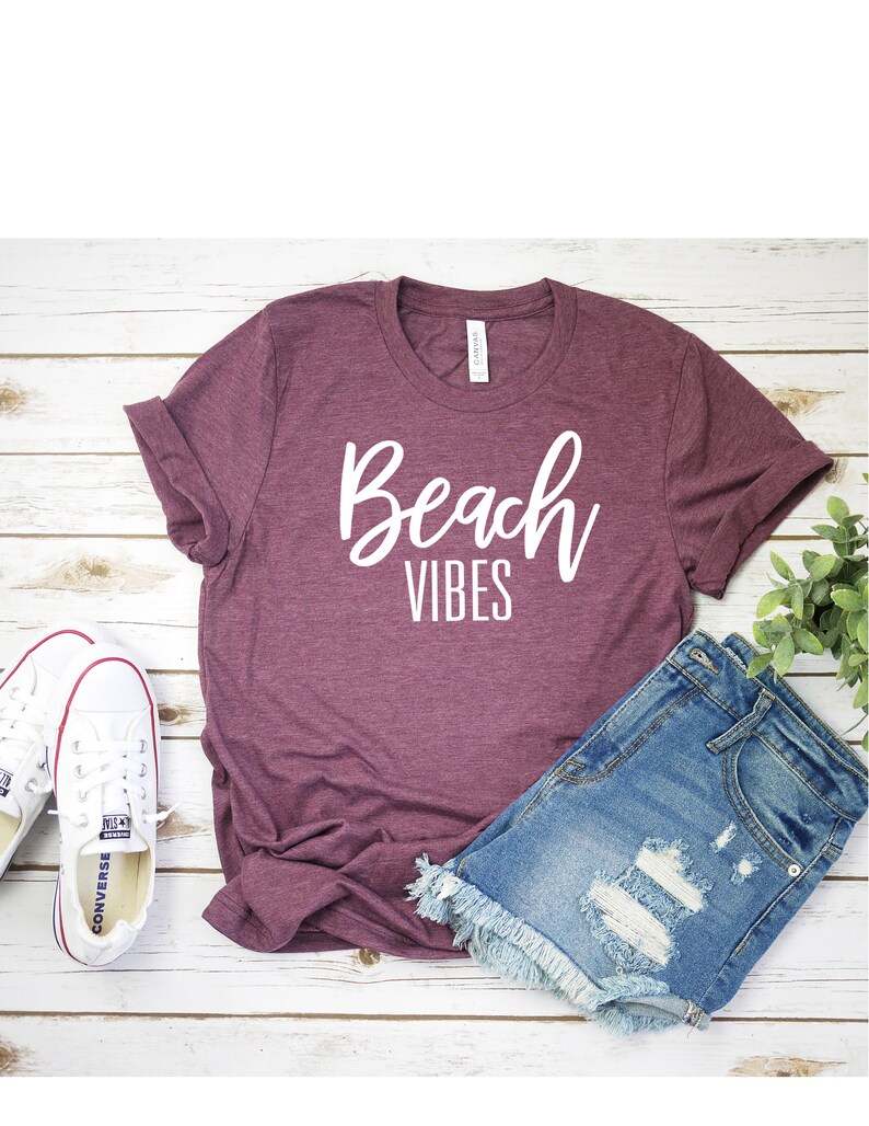 K&ouml;nnte beinhalten: Ein maroonfarbenes T-Shirt mit dem Text "Beach Vibes" in wei&szlig;en Buchstaben. Das Shirt liegt flach auf einer wei&szlig;en Holzoberfl&auml;che mit einem Paar wei&szlig;en Turnschuhen und einer Jeans-Shorts.