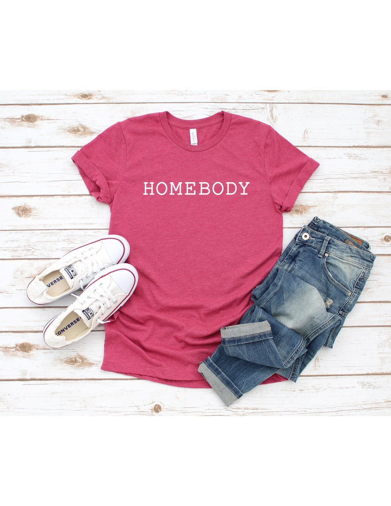 Pode incluir: Uma camiseta rosa com o texto "HOMEBODY" estampado em branco na frente. A camiseta est&aacute; colocada sobre uma superf&iacute;cie de madeira branca com um par de t&ecirc;nis brancos e uma cal&ccedil;a jeans azul.