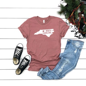 May include: A mauve t-shirt with a white outline of the state of North Carolina and the text "NORTH CAROLINA" with a heart replacing the "O" in "NORTH".