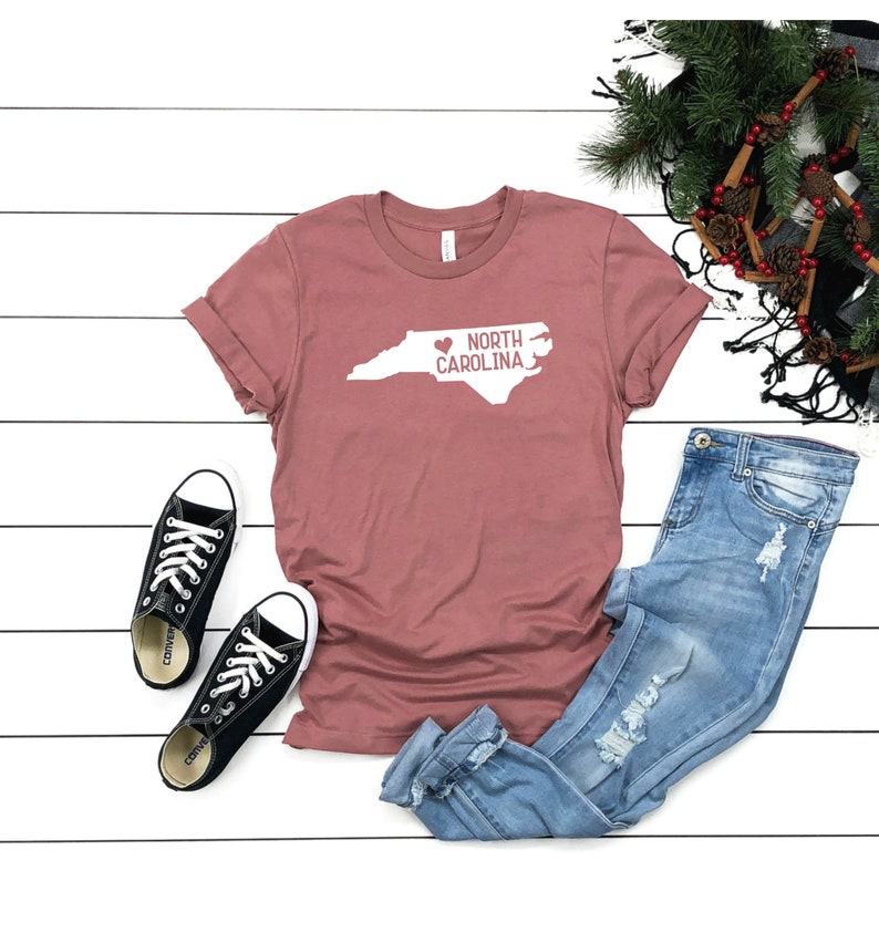 May include: A mauve t-shirt with a white outline of the state of North Carolina and the text "NORTH CAROLINA" with a heart replacing the "O" in "NORTH".