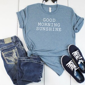 May include: A blue t-shirt with the text "GOOD MORNING SUNSHINE" in white. The shirt is paired with blue jeans and navy blue sneakers.