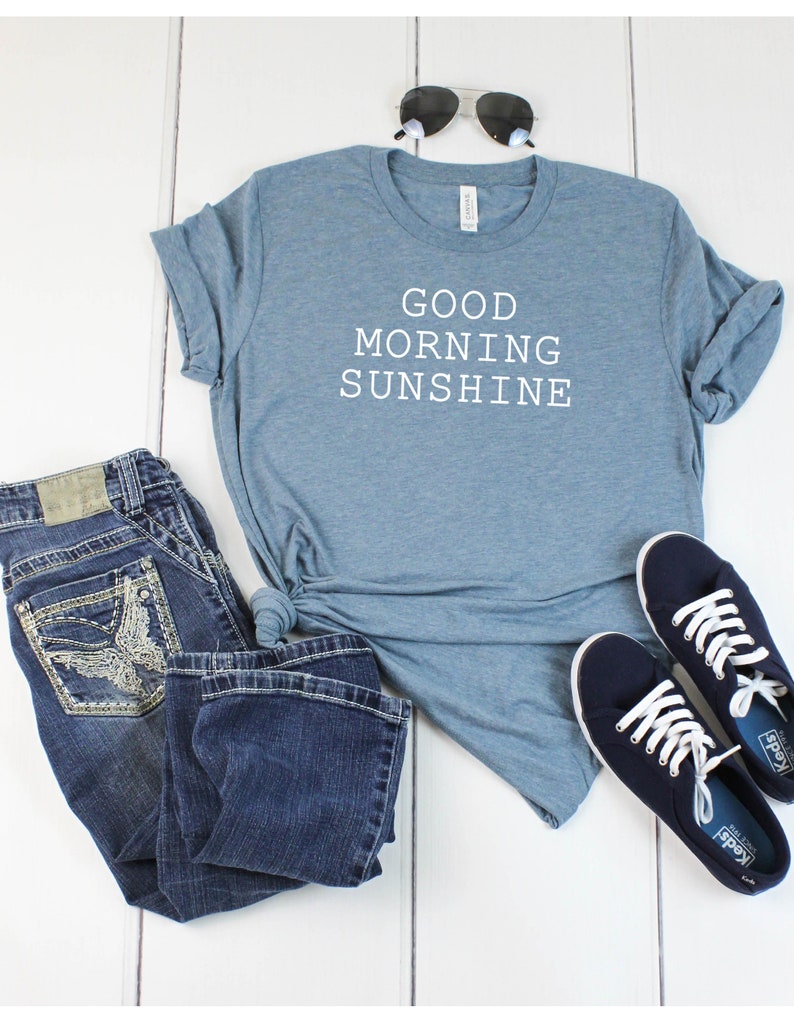 May include: A blue t-shirt with the text "GOOD MORNING SUNSHINE" in white. The shirt is paired with blue jeans and navy blue sneakers.