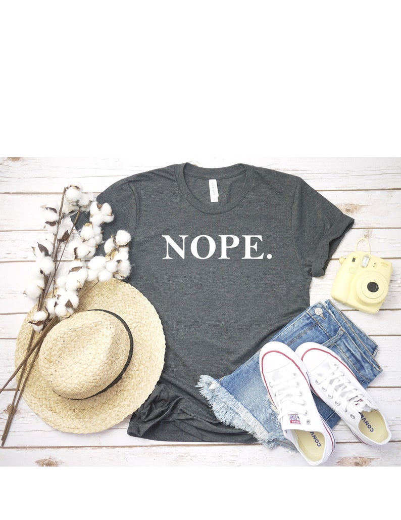 May include: A dark gray t-shirt with the word "NOPE." printed in white on the front. The shirt is laid flat on a wooden surface with a straw hat, denim shorts, white sneakers, and a yellow camera.