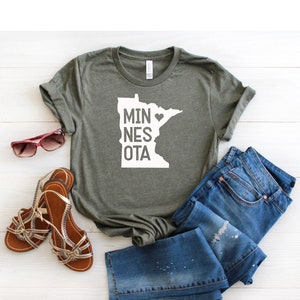 May include: A green t-shirt with a white outline of the state of Minnesota and the text "MINNESOTA" with a heart replacing the "O". The shirt is laid flat on a white wooden surface with a pair of brown sandals and blue jeans.