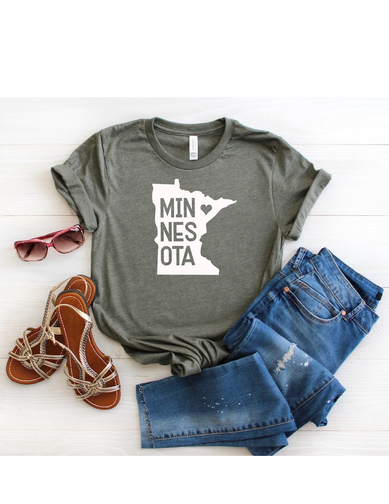 May include: A green t-shirt with a white outline of the state of Minnesota and the text "MINNESOTA" with a heart replacing the "O". The shirt is laid flat on a white wooden surface with a pair of brown sandals and blue jeans.
