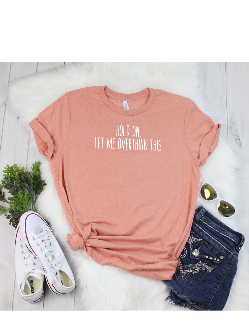May include: A peach colored t-shirt with the text "HOLD ON, LET ME OVERTHINK THIS" printed in white.
