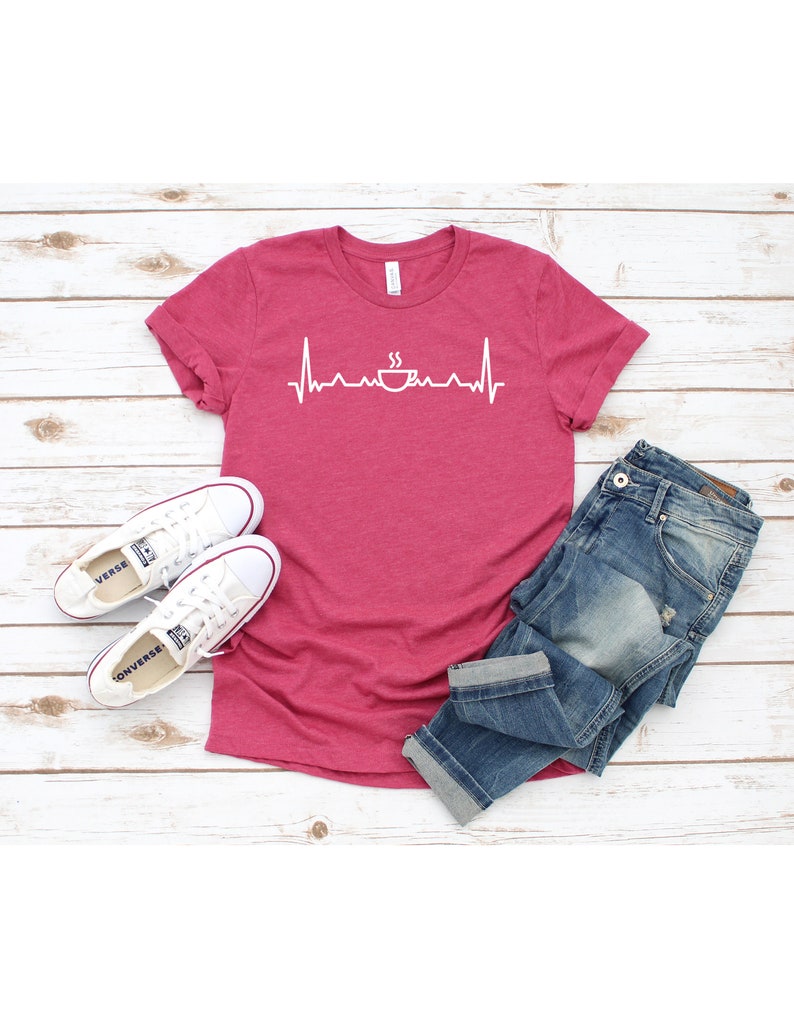 Puede incluir: Una camiseta rosa con un gr&aacute;fico blanco de una taza de caf&eacute; con vapor saliendo de ella y una l&iacute;nea de latido del coraz&oacute;n. La camiseta est&aacute; sobre una superficie de madera blanca con un par de zapatillas blancas y unos vaqueros azules.