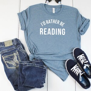 May include: A blue t-shirt with the text "I'd rather be reading" in white. The shirt is paired with blue jeans and navy blue sneakers.