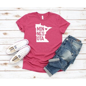 May include: A pink t-shirt with a white outline of the state of Minnesota and the text "MIN&hearts;NESOTA". The shirt is laid out on a white wooden surface with a pair of white sneakers and blue jeans.