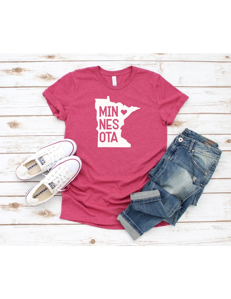 May include: A pink t-shirt with a white outline of the state of Minnesota and the text "MIN&hearts;NESOTA". The shirt is laid out on a white wooden surface with a pair of white sneakers and blue jeans.