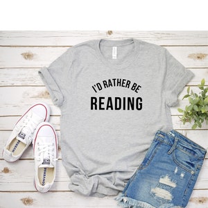 May include: A gray t-shirt with the text "I'd rather be reading" in black lettering. The shirt is laid flat on a wooden surface with a pair of white sneakers and denim shorts.