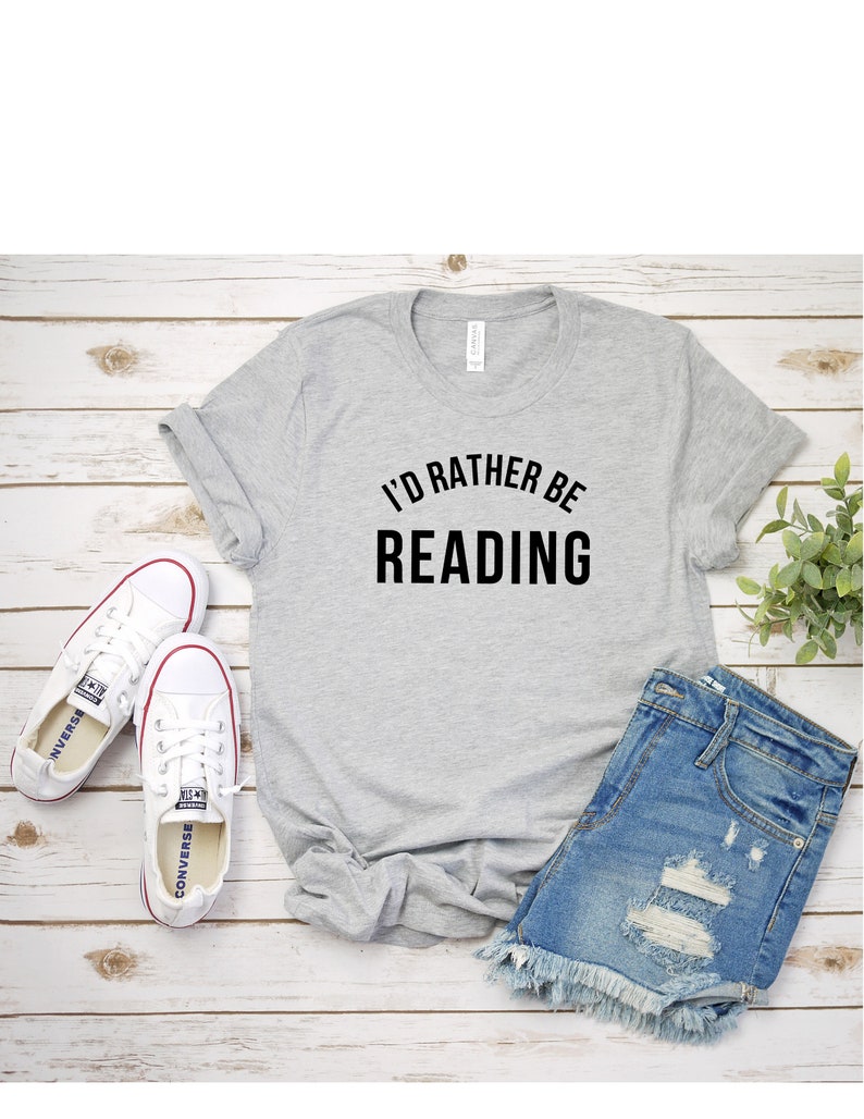 May include: A gray t-shirt with the text "I'd rather be reading" in black lettering. The shirt is laid flat on a wooden surface with a pair of white sneakers and denim shorts.