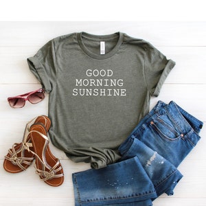 May include: A green t-shirt with the text "GOOD MORNING SUNSHINE" printed in white on the front. The t-shirt is paired with blue jeans and sandals.