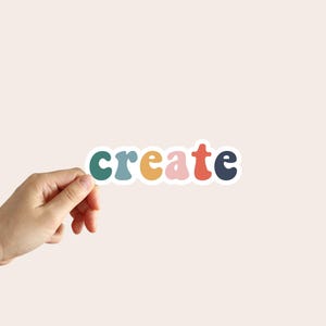 May include: A colorful sticker with the word "create" in a retro font. The word is composed of various colors, including teal, blue, yellow, pink, and red. The sticker is held by a hand against a neutral background.