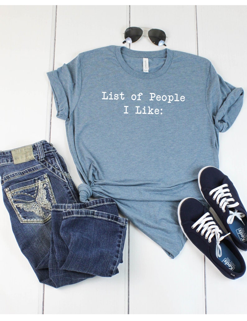 May include: A blue t-shirt with the text "List of People I Like:" printed in white on the front. The shirt is laid out on a white wooden surface with a pair of sunglasses, blue jeans, and a pair of blue sneakers.