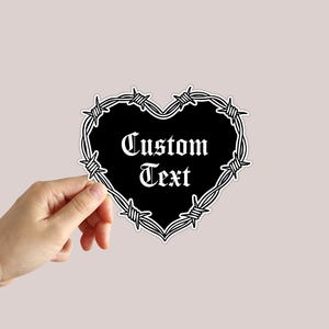May include: A heart-shaped sticker featuring a black center surrounded by a white barbed wire border. The words Custom Text are printed in the center in a white gothic-style font. The sticker has a thin white outline around the entire shape.