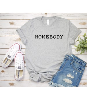 Pode incluir: Uma camiseta cinza com o texto "HOMEBODY" estampado na frente. A camiseta est&aacute; colocada sobre uma superf&iacute;cie de madeira branca com um par de t&ecirc;nis brancos e shorts jeans.