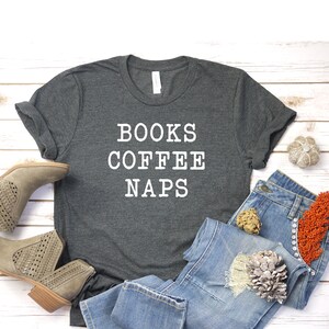 May include: A dark gray t-shirt with the text "BOOKS COFFEE NAPS" printed in white. The shirt is laid flat on a white wooden surface with a pair of brown ankle boots, blue jeans, and decorative items.