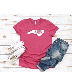 May include: A pink t-shirt with a white outline of the state of North Carolina and a heart over the state. The text "NORTH CAROLINA" is written inside the state outline. The t-shirt is laid out on a white wooden surface with a pair of white sneakers and blue jeans.