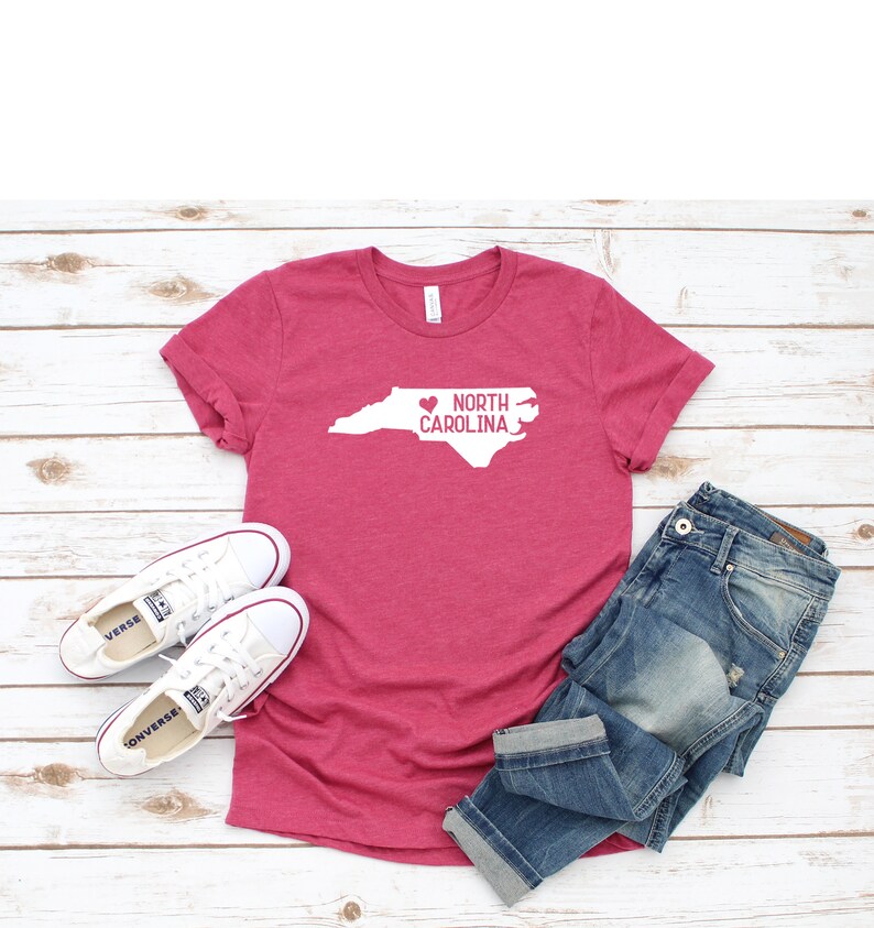 May include: A pink t-shirt with a white outline of the state of North Carolina and a heart over the state. The text "NORTH CAROLINA" is written inside the state outline. The t-shirt is laid out on a white wooden surface with a pair of white sneakers and blue jeans.