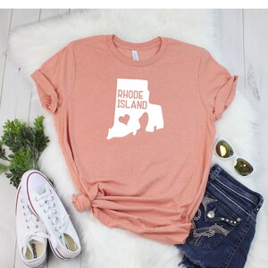 K&ouml;nnte beinhalten: Ein pfirsichfarbenes T-Shirt mit einer wei&szlig;en Umrandung des Bundesstaates Rhode Island und dem Text "Rhode Island" mit einem kleinen Herzen innerhalb der Bundesstaatenumrandung.