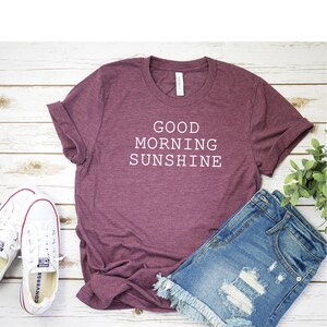 May include: A maroon t-shirt with the text "GOOD MORNING SUNSHINE" in white. The shirt is laid flat on a white wooden surface with a pair of white sneakers and denim shorts.