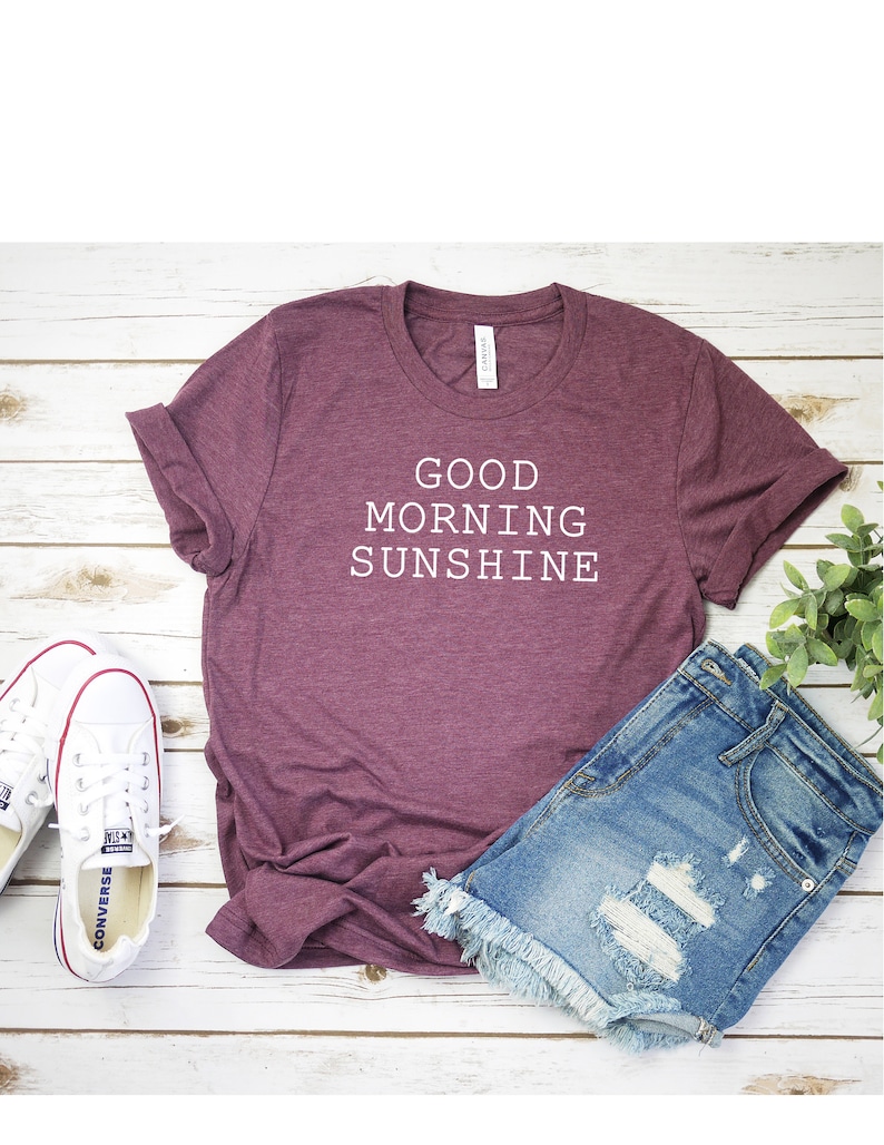 May include: A maroon t-shirt with the text "GOOD MORNING SUNSHINE" in white. The shirt is laid flat on a white wooden surface with a pair of white sneakers and denim shorts.