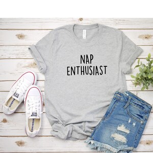 May include: A gray t-shirt with the text "NAP ENTHUSIAST" printed in black. The shirt is laid flat on a white wooden surface with a pair of white sneakers and denim shorts.
