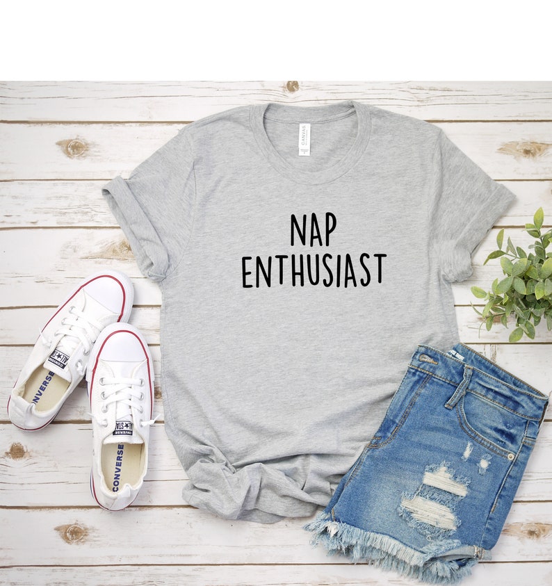 May include: A gray t-shirt with the text "NAP ENTHUSIAST" printed in black. The shirt is laid flat on a white wooden surface with a pair of white sneakers and denim shorts.