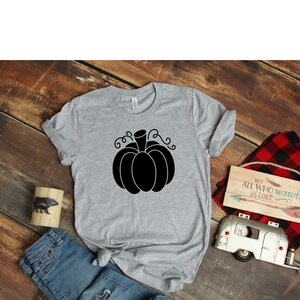 May include: A gray t-shirt with a black pumpkin graphic. The pumpkin has a stem and curly vines. The shirt is on a wooden surface with a pair of blue jeans, a wooden sign, and a toy camper.