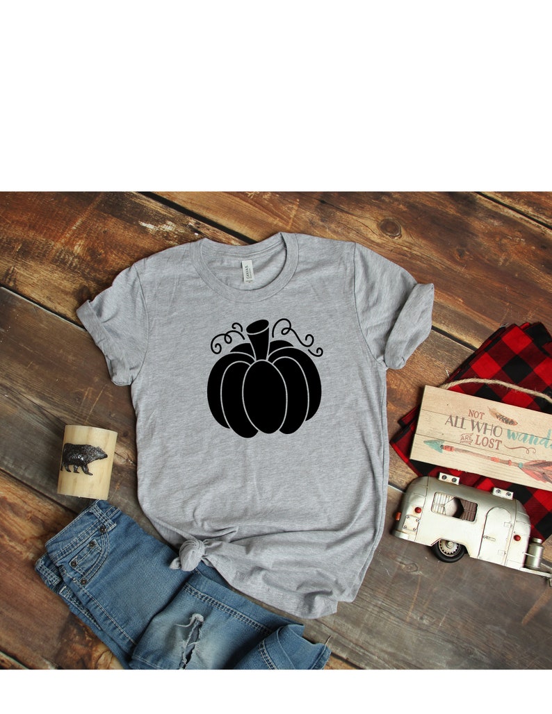 May include: A gray t-shirt with a black pumpkin graphic. The pumpkin has a stem and curly vines. The shirt is on a wooden surface with a pair of blue jeans, a wooden sign, and a toy camper.