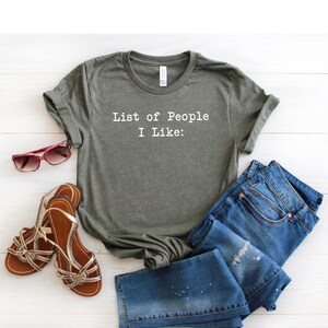 May include: A green t-shirt with the text "List of People I Like:" printed on the front. The shirt is laid out on a white wooden surface with a pair of sandals and blue jeans.