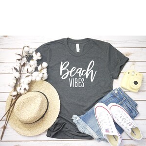 K&ouml;nnte beinhalten: Ein graues T-Shirt mit dem Text "Beach Vibes" in wei&szlig;en Buchstaben. Das Shirt liegt auf einer wei&szlig;en Holzoberfl&auml;che mit einem Strohhut, Jeans-Shorts, wei&szlig;en Turnschuhen und einer gelben Kamera.