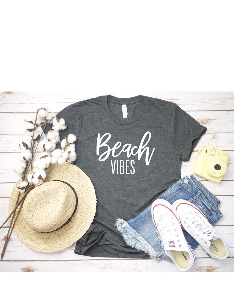 K&ouml;nnte beinhalten: Ein graues T-Shirt mit dem Text "Beach Vibes" in wei&szlig;en Buchstaben. Das Shirt liegt auf einer wei&szlig;en Holzoberfl&auml;che mit einem Strohhut, Jeans-Shorts, wei&szlig;en Turnschuhen und einer gelben Kamera.