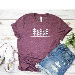 May include: A maroon t-shirt with a white graphic of a row of five stylized pine trees. The t-shirt is laying on a white wooden surface with a pair of white sneakers and a pair of blue denim shorts.