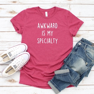 May include: A pink t-shirt with the text "Awkward is my specialty" in white lettering. The shirt is paired with blue jeans and white sneakers.