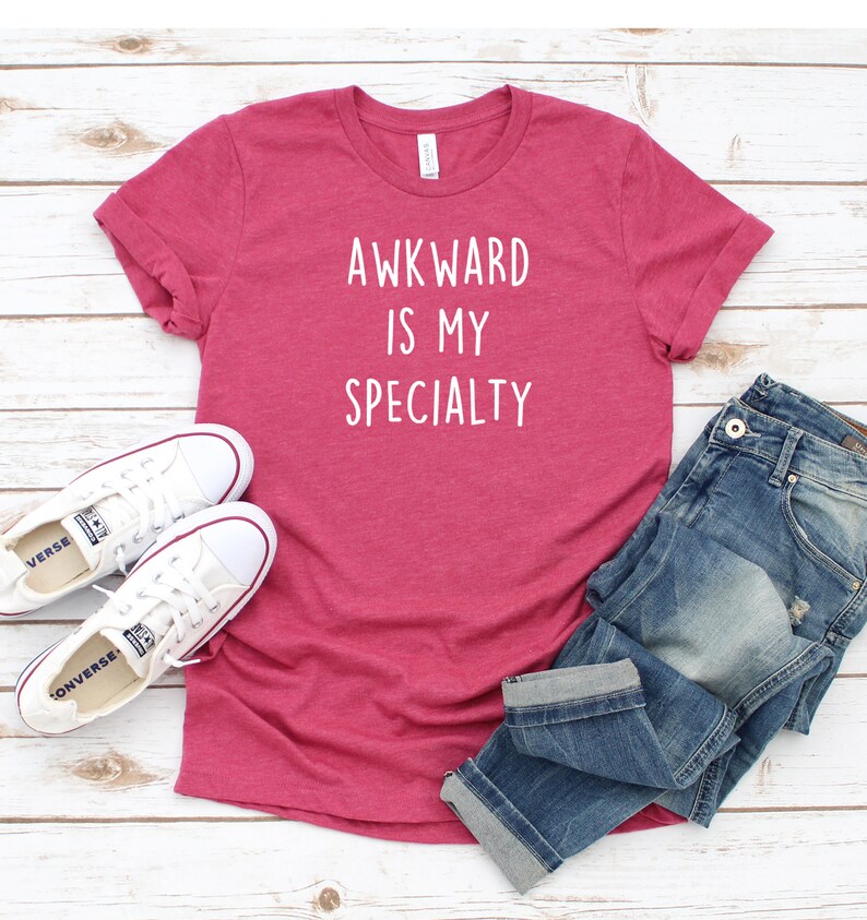 May include: A pink t-shirt with the text "Awkward is my specialty" in white lettering. The shirt is paired with blue jeans and white sneakers.