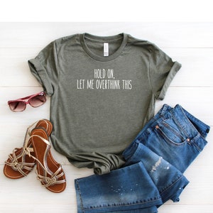 May include: A green t-shirt with the text "Hold on, let me overthink this" printed on the front. The shirt is laid out on a white wooden surface with a pair of sandals and blue jeans.