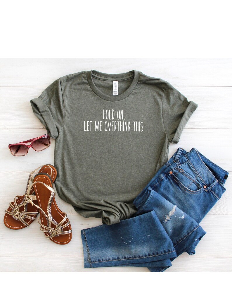 May include: A green t-shirt with the text "Hold on, let me overthink this" printed on the front. The shirt is laid out on a white wooden surface with a pair of sandals and blue jeans.