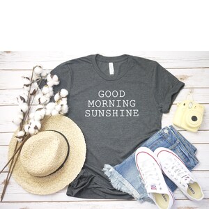 May include: A dark gray t-shirt with the text "GOOD MORNING SUNSHINE" in white lettering. The shirt is laid flat on a white wooden surface with a straw hat, denim shorts, white sneakers, and a yellow camera.