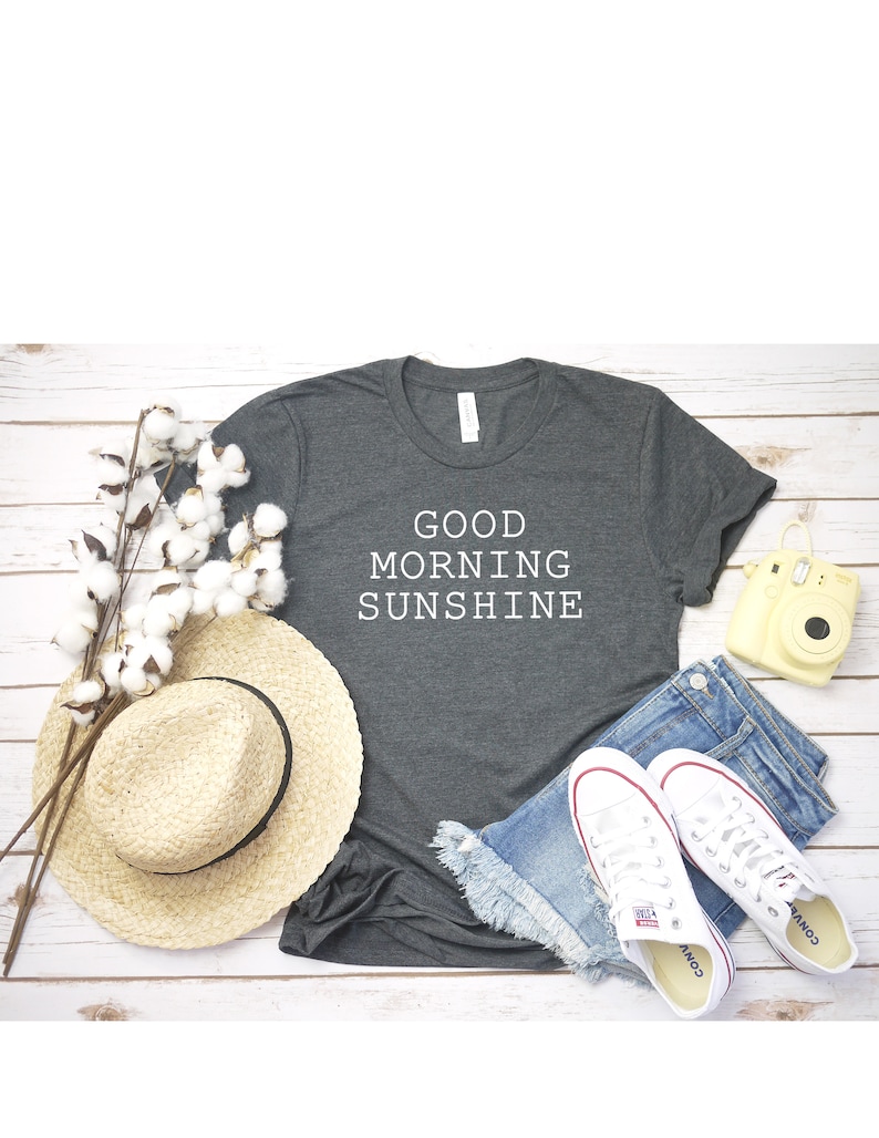 May include: A dark gray t-shirt with the text "GOOD MORNING SUNSHINE" in white lettering. The shirt is laid flat on a white wooden surface with a straw hat, denim shorts, white sneakers, and a yellow camera.