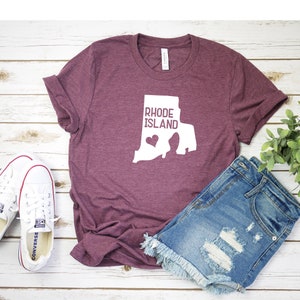 K&ouml;nnte beinhalten: Ein maroonfarbenes T-Shirt mit einer wei&szlig;en Umrandung des Bundesstaates Rhode Island und dem Text "Rhode Island" innerhalb der Umrandung. Ein kleines wei&szlig;es Herz befindet sich innerhalb der Umrandung. Das T-Shirt liegt auf einer wei&szlig;en Holzoberfl&auml;che mit einem Paar wei&szlig;en Turnschuhen und einer hellblauen Jeans-Shorts.