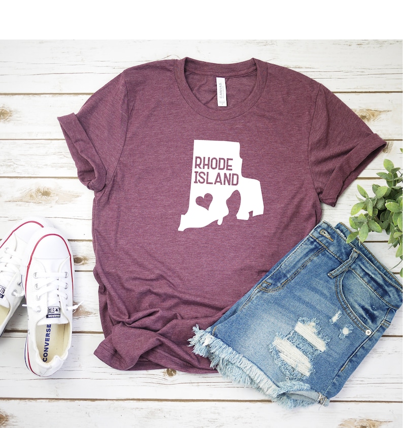 K&ouml;nnte beinhalten: Ein maroonfarbenes T-Shirt mit einer wei&szlig;en Umrandung des Bundesstaates Rhode Island und dem Text "Rhode Island" innerhalb der Umrandung. Ein kleines wei&szlig;es Herz befindet sich innerhalb der Umrandung. Das T-Shirt liegt auf einer wei&szlig;en Holzoberfl&auml;che mit einem Paar wei&szlig;en Turnschuhen und einer hellblauen Jeans-Shorts.
