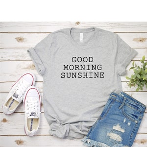 May include: Gray t-shirt with the text "GOOD MORNING SUNSHINE" printed on it. The t-shirt is laid out on a wooden surface with a pair of white sneakers and denim shorts.