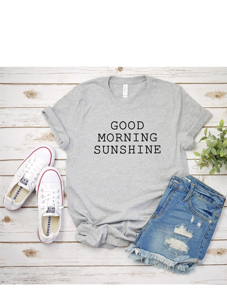 May include: Gray t-shirt with the text "GOOD MORNING SUNSHINE" printed on it. The t-shirt is laid out on a wooden surface with a pair of white sneakers and denim shorts.