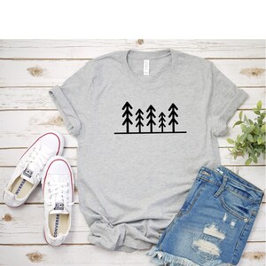 May include: A gray t-shirt with a black graphic of five stylized pine trees. The t-shirt is paired with denim shorts and white sneakers.