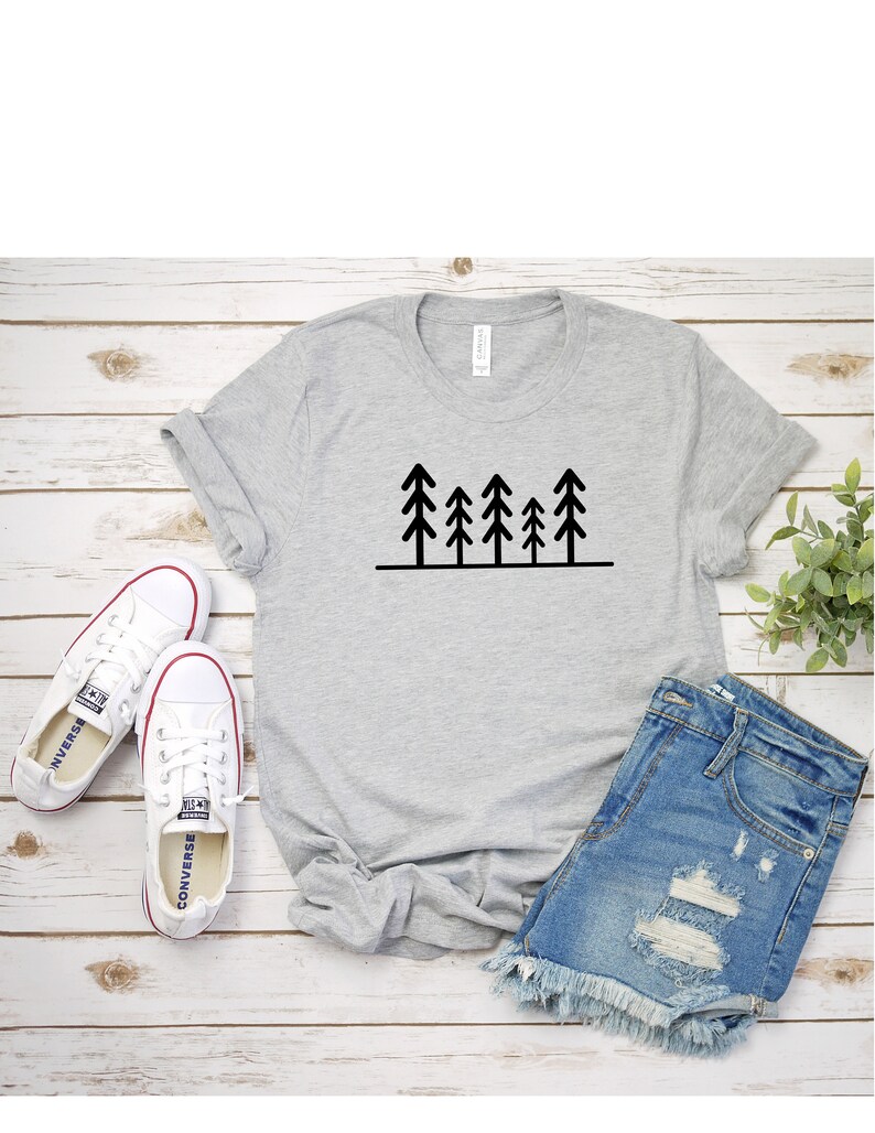 May include: A gray t-shirt with a black graphic of five stylized pine trees. The t-shirt is paired with denim shorts and white sneakers.