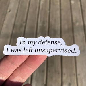May include: A white sticker with the text "In my defense, I was left unsupervised." The sticker has a rounded rectangular shape and is held by a hand against a wooden background.