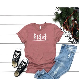 May include: A mauve t-shirt with a white graphic of five stylized trees. The t-shirt is paired with light blue jeans and black sneakers.