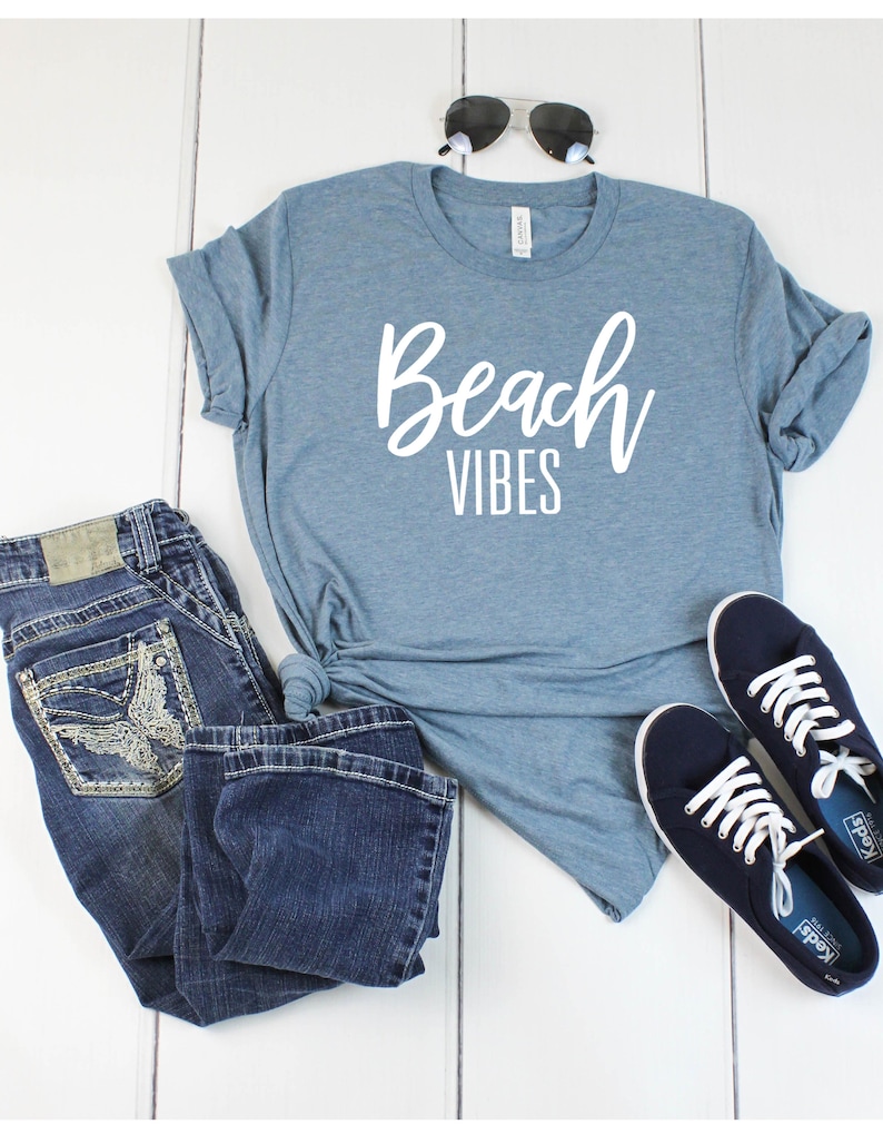 K&ouml;nnte beinhalten: Ein blaues T-Shirt mit dem Schriftzug "Beach Vibes" in wei&szlig;en Buchstaben. Das Shirt wird mit einer blauen Jeans und dunkelblauen Sneakers kombiniert.
